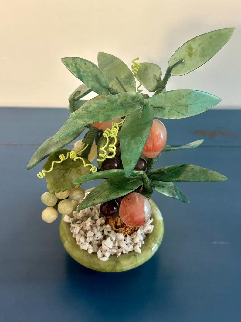 May include: A small, decorative jade tree with green leaves, red-orange fruit, and clusters of grapes. The tree is potted in a round, green container filled with small white stones. The background is a blue surface.