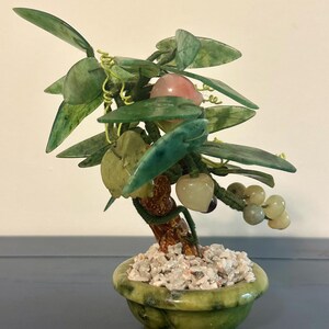 May include: A decorative jade bonsai tree in a green ceramic pot. The tree features green leaves, a brown trunk, and various stone fruits in shades of pink, white, and green. The pot is filled with small white stones.