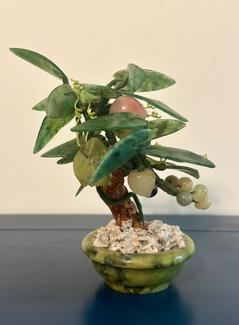 May include: A decorative jade bonsai tree in a green ceramic pot. The tree features green leaves, a brown trunk, and various stone fruits in shades of pink, white, and green. The pot is filled with small white stones.