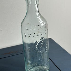 May include: An antique, clear glass bottle with the embossed text "SCOTTS EMULSION" and a figure of a man. The bottle has a rectangular shape with rounded shoulders and a narrow neck. The bottle is on a blue surface.