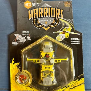 May include: A yellow and gray Hexbug Warriors Battling Robots toy, packaged in a black and yellow blister pack. The package includes the text "TRONIKON" and "REAL RUNNING MOTORS". The toy is recommended for ages 6 and up.