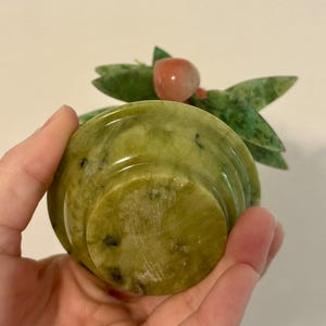 May include: A carved jade sculpture of a small bowl with a fruit and leaf design. The bowl is a light green color with darker green and black flecks. The fruit is a reddish-orange color, and the leaves are green. The sculpture is held in a hand.