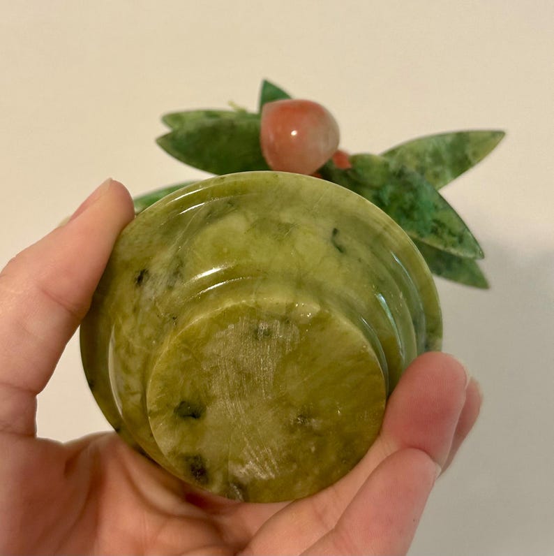 May include: A carved jade sculpture of a small bowl with a fruit and leaf design. The bowl is a light green color with darker green and black flecks. The fruit is a reddish-orange color, and the leaves are green. The sculpture is held in a hand.