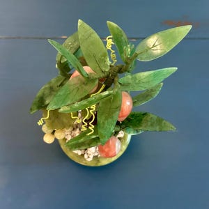 May include: A decorative jade tree with green leaves and red-orange fruit. The tree is in a green pot filled with small white stones. Yellow wire accents the branches, creating a unique and artistic display.