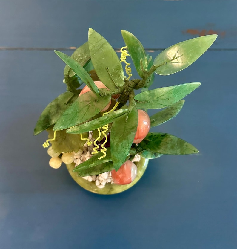 May include: A decorative jade tree with green leaves and red-orange fruit. The tree is in a green pot filled with small white stones. Yellow wire accents the branches, creating a unique and artistic display.