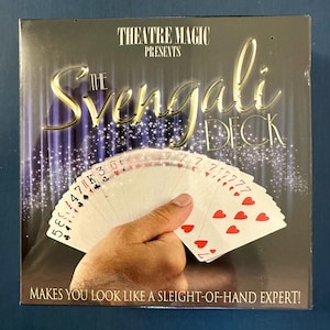 May include: A box for "The Svengali Deck" magic trick. The box is dark with gold lettering and a hand holding a fanned deck of cards. The cards are white with red hearts and black clubs. The text reads "MAKES YOU LOOK LIKE A SLEIGHT-OF-HAND EXPERT!"