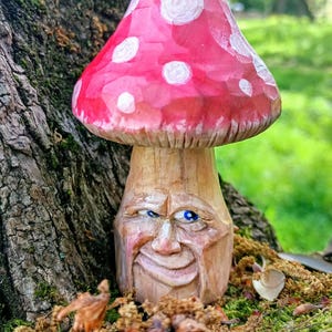 Puede incluir: Una escultura de hongo de madera tallada a mano. El sombrero del hongo está pintado de rojo brillante con lunares blancos. El tallo presenta una cara tallada con ojos azules y una expresión sonriente. El fondo es natural.