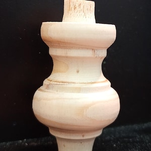 May include: A turned wooden furniture leg, unfinished and ready for staining or painting. The leg features a bulbous lower section, a central hourglass shape, and a cylindrical top. The wood grain is visible, showcasing the natural texture of the material.