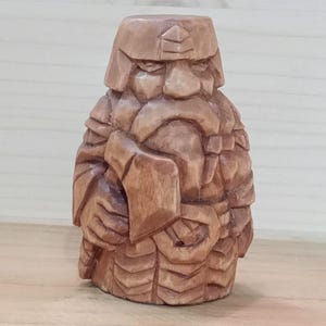 Könnte beinhalten: Handgeschnitzte Holzfigur eines bärtigen Zwergs in Rüstung. Die Figur ist braun mit detaillierten Schnitzereien, die einen Helm, Bart und Tunika darstellen. Der Zwerg hält eine kleine Axt. Die Figur ist ein dekorativer Gegenstand.