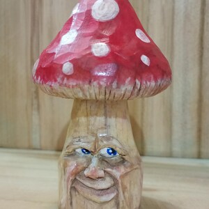 May include: Hand-carved wooden mushroom sculpture with a red cap and white spots. The mushroom has a detailed face carved into the stem, featuring blue eyes and a smiling expression. The sculpture is set against a wooden background.