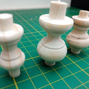 Könnte beinhalten: Drei unfertige Holzmöbelfüße, jeweils mit einem einzigartigen gedrehten Design. Das helle Holz zeigt die natürliche Maserung. Sie liegen auf einer grünen Schneidematte mit Rastermuster, was auf DIY-Projekte hindeutet.