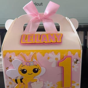 May include: A white gift box with a pink bow, featuring a bee-themed design. The box has a yellow and pink color scheme, with the name "LEILANY" and the number "1" on the front. A cartoon bee is also featured.
