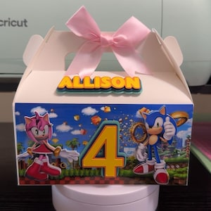 May include: A white party favor box decorated with Sonic the Hedgehog characters and the number 4. The box has a pink bow on top and the name "ALLISON" in yellow letters. The box is on a white rotating stand.