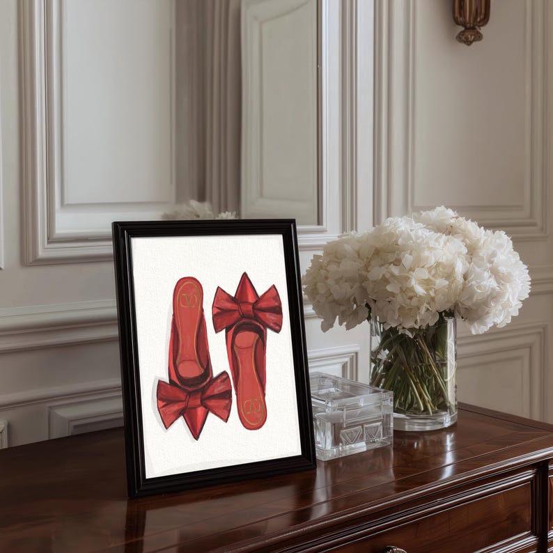 Op de afbeelding: Een ingelijste print met rode ballerina's met strikaccenten. Het kunstwerk staat op een donker houten oppervlak, met een vaas met witte bloemen en een doorzichtige glazen doos. De lijst is zwart.