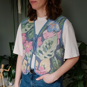 Può includere: Un gilet con stampa floreale con finiture blu e chiusura con lacci. Il gilet presenta un motivo colorato di fiori e foglie rosa, blu e verdi su sfondo blu scuro. Il gilet è indossato sopra una maglietta bianca e jeans blu.