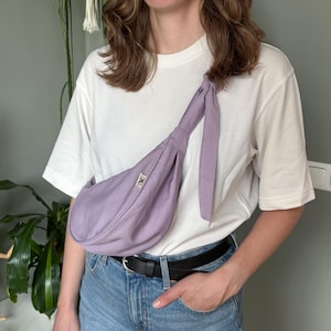 Puede incluir: Un bolso bandolera de color lavanda con una correa larga atada con un nudo. El bolso se lleva sobre el hombro y cruzado por el cuerpo. El bolso tiene una pequeña etiqueta rectangular con un logotipo en la parte delantera. La persona lleva una camiseta blanca y vaqueros azules.