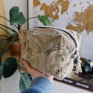 Op de afbeelding: Een beige en bruin patroon make-up tasje met een zwarte rits en een klein label. De tas wordt door een persoon vastgehouden. De achtergrond bevat een plant en een wereldkaart.