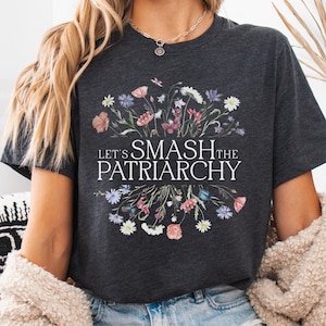 Könnte beinhalten: Dunkelgraues T-Shirt mit floralem Design und dem Text "Let's Smash the Patriarchy" in Weiß. Das Blumenarrangement umfasst rosa, lila und weiße Blumen. Das T-Shirt wird mit Jeansshorts und einer cremefarbenen Strickjacke getragen.