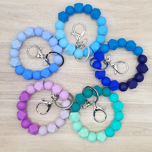 May include: Five silicone wristlet keychains in shades of blue, purple, and teal. Each keychain features a variety of geometric and round beads and a silver-colored clasp. The keychains are arranged on a light wood-grain surface.