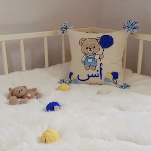 May include: A cream cushion featuring an embroidered teddy bear holding a blue balloon. The bear wears blue dungarees. The cushion has blue and yellow pom-poms and Arabic text. A small teddy bear and yarn balls rest on a white fluffy surface.