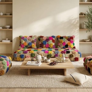 Może przedstawiać: Niska sofa i dwie pasujące kwadratowe poduszki podłogowe wykonane z naturalnej beżowej tkaniny, ozdobione siatką kolorowych, teksturowanych pomponów w odcieniach różu, błękitu, żółci i zieleni. Do jednej z poduszek przymocowana jest mała metka.