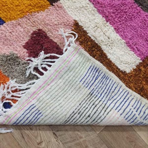 May include: A colorful, geometric rug with a mix of textures and hues. The rug features a patchwork design with sections of pink, orange, brown, and white. The edge of the rug is visible, showing a striped pattern with fringe.