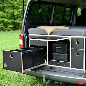 Könnte beinhalten: Ein maßgefertigter Camper-Van-Innenraum mit Bett, Schubladen und eingebauter Kühlbox. Die Schubladen sind schwarz mit strukturierter Oberfläche, und der Innenraum hat eine neutrale Farbpalette. Der Van steht auf Gras.