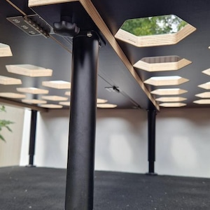 Könnte beinhalten: Verstellbarer Tisch in Schwarz und Holzoptik mit geometrischem Design. Die Tischplatte hat sechseckige Aussparungen. Der Tisch hat schwarze, zylindrische Beine und höhenverstellbare Funktionen. Konzipiert für Vielseitigkeit und Funktionalität.