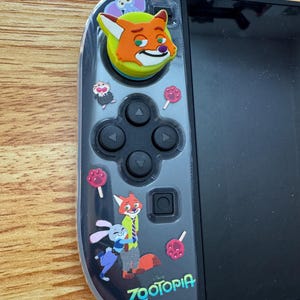 以下が含まれることがあります： 黒と透明のNintendo Switch Joy-Conコントローラー、ズートピアをテーマにしたスキン付き。スキンには、ニック・ワイルド、ジュディ・ホップス、その他のキャラクターの漫画画像、ズートピアのロゴ、ピンクのロリポップが描かれています。