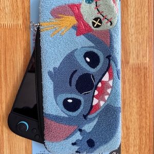 以下が含まれることがあります： ディズニーの「リロ＆スティッチ」に登場するスティッチのキャラクターが描かれた青いぬいぐるみケース。ケースにはジッパー開閉とリストストラップが付いています。デザインにはスティッチの顔と人形のようなキャラクターが含まれています。黒いゲーム機が一部見えています。
