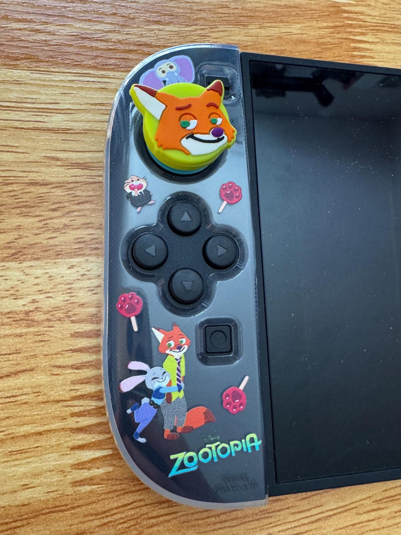 以下が含まれることがあります： 黒と透明のNintendo Switch Joy-Conコントローラー、ズートピアをテーマにしたスキン付き。スキンには、ニック・ワイルド、ジュディ・ホップス、その他のキャラクターの漫画画像、ズートピアのロゴ、ピンクのロリポップが描かれています。