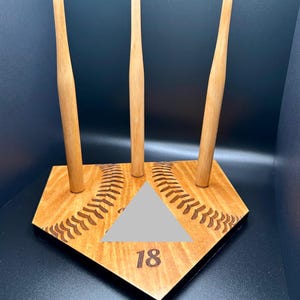 May include: A wooden baseball bat display stand. The stand features three wooden bat holders with pink tips, set on a baseball home plate design. The home plate has a grey triangle and the number 18.
