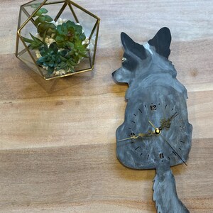 Könnte beinhalten: Dekorative Wanduhr in Hundeform, bemalt in Grau- und Weißtönen. Die Uhr hat goldfarbene Zeiger und einen herabhängenden Schwanz. Links befindet sich ein geometrisches Glas-Terrarium mit Grünpflanzen.