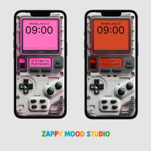 May include: Two phone cases with a retro video game design. One case has a pink screen displaying "Monday, June 23, 09:00" and "IT'S TIME TO PLAY GAMES." The other case has an orange screen with the same text. The cases have a white and grey colour scheme with black accents.