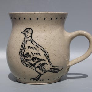 May include: A beige ceramic mug with a speckled finish and a rounded body. The mug features a black line drawing of a bird on the front, with a row of small black dots encircling the top rim and the base. It has a curved handle on the right side.
