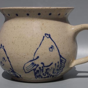 May include: A round, speckled beige ceramic mug featuring a blue hand-painted fish design on the exterior. The mug has a wide, flared rim decorated with a series of small blue dots and a sturdy, curved handle on the right side of the vessel.