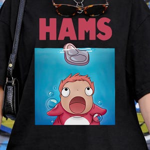 May include: Black t-shirt featuring the word "HAMS" in red above a cartoon image of a character from the animated film "Ponyo". The character is underwater, looking upwards with a surprised expression.