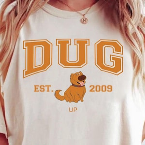 Può includere: T-shirt color avorio con la scritta "DUG" a blocchi arancioni, il testo "EST. 2009" e un'illustrazione di un cane dei cartoni animati. La parola "UP" è sotto il cane. La maglietta è in stile casual.