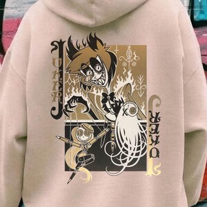 May include: Beige hoodie with a graphic print featuring a cartoon character with flames, a doll, and the text "Joker". The design is in shades of brown, black, and gold. The hoodie has a hood and long sleeves.