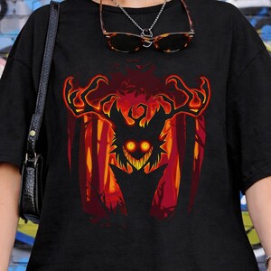 May include: Black t-shirt featuring a vibrant graphic of a horned creature with glowing yellow eyes, set against a fiery forest backdrop. The design is bold and eye-catching, perfect for fans of fantasy or dark art.