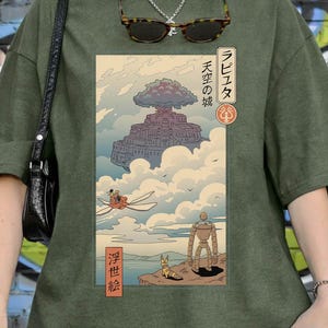 May include: A moss green Comfort Colors t-shirt with a Japanese anime-style illustration. The graphic shows a floating castle, a flying machine, and a robot. The text on the shirt includes Japanese characters and the words "Comfort Colors".