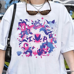 May include: White t-shirt featuring a colorful graphic design with cartoon characters in pink, blue, and purple. The design includes the letters "I" and "P" within a circular pattern. The shirt has short sleeves and a casual fit.