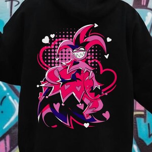 May include: Black hoodie featuring a vibrant cartoon jester design on the back. The jester is depicted in shades of pink and red, with a mischievous grin and heart-shaped accents. The hoodie has a drawstring hood and long sleeves.