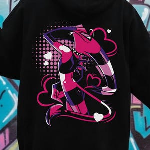 May include: Black hoodie featuring a vibrant graphic on the back. The design includes a stylized image with pink, purple, and black elements, accented by hearts and a dotted background. The hoodie has a hood and long sleeves.