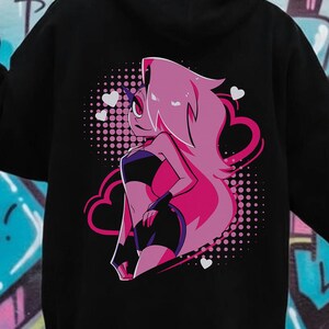 May include: Black hoodie featuring a pink and purple anime-style graphic on the back. The graphic depicts a female character with pink hair, a black top, and shorts, set against a pink dotted background with heart accents. The hoodie is worn by a person in front of a graffiti wall.