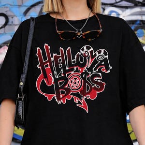 May include: Black t-shirt with the text "Helluva Boss" in red and white stylized lettering. The design includes a pentagram and other graphic elements. The shirt is worn by a person in front of a graffiti wall.
