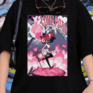 May include: Black t-shirt featuring a colorful graphic of a character with the text "LOVE POTION" and "BY VELVET CUTE". The graphic includes pink hearts and a pink cloud-like base.