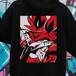 May include: Black hoodie featuring a red and white graphic of a fox-like character on the back. The design is a stylized illustration with sharp lines and bold colors. The hoodie has a hood and long sleeves.