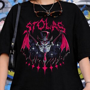 May include: Black t-shirt featuring a graphic of a bat-like creature with red wings, a top hat, and the word "STOLAS" in pink. The design includes stars and a dark, cosmic background. The shirt is worn by a person.