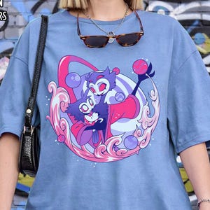 May include: A light blue t-shirt with a graphic of two cartoon characters in pink and purple, surrounded by a pink and white swirl design. The shirt has the text "BLUE JEAN COMFORT COLORS" at the top.